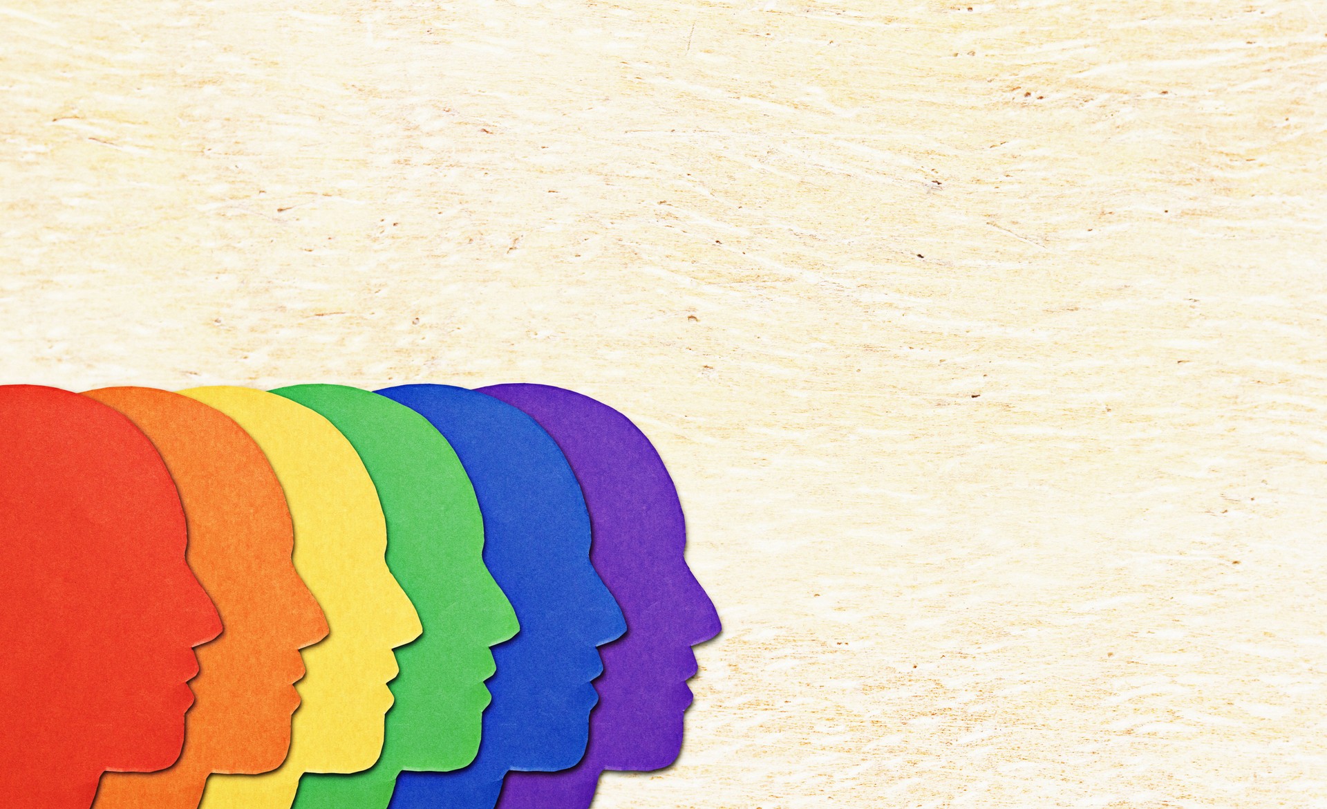 Cutout faces in the colors of gay pride, overlapping, on a textured wood background with copy space